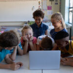 Front view of a diverse group of eight schoolchildren gathered around desks using a laptop computer together in an elementary school classroom, talking and laughing together