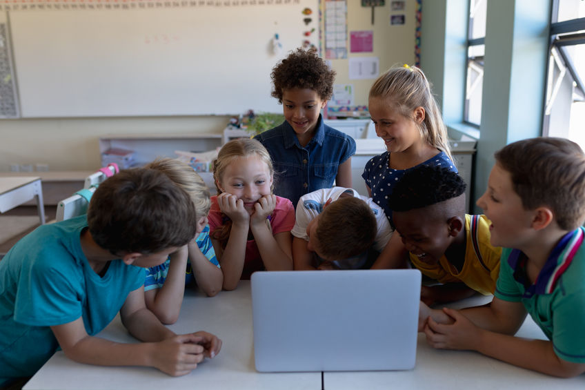Front view of a diverse group of eight schoolchildren gathered around desks using a laptop computer together in an elementary school classroom, talking and laughing together